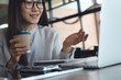 © tippapatt - Smiling asian businesswoman holding coffee cup during video call on laptop in cafe, remote work, freelance at work, business casual lifestyle concept.