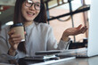 © tippapatt - Smiling asian businesswoman holding coffee cup during video call on laptop in cafe, remote work, freelance at work, business casual lifestyle concept.