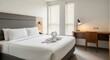 © RANJIT - Hotel bedroom interior with a gray upholstered headboard, white bedding, swan towels, wooden desk, chair, and blinds on the windows