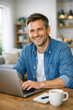 © Oleg - Man wearing casual attire and smiling at the camera while using a laptop in his home office setting.