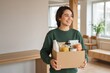 © MrHamster - A young woman smiles happily while carrying a cardboard box filled with groceries in her new apartment.