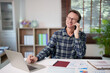 © Tj - Mature man working from home, smiling while talking on a smartphone and taking notes on paper, managing business finances and communicating remotely