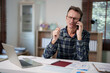 © Tj - Mature businessman talking on a mobile phone with a concerned expression. Holding a pen and managing paperwork and a laptop at his home office desk