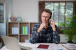 © Tj - Middle-aged man talking on a mobile phone while working from home, thinking and analyzing business data, concentrating on a remote work call, solving problems and making decisions