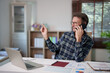 © Tj - Mature man working from his home office, talking on a mobile phone while pointing with a pen, surrounded by a laptop, documents, and financial charts on his desk