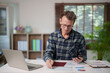 © Tj - Senior man analyzing financial reports and calculating household budget, managing personal finance with paperwork, smartphone, and laptop in a home office setting