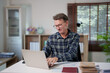 © Tj - Happy mature man in a plaid shirt and glasses using a laptop at a white desk in his home office, enjoying remote work and professional success