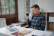 © Tj - Senior man wearing glasses and a plaid shirt, working on his laptop at a desk in a home office environment, holding a cup of coffee and reviewing documents