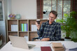 © Tj - Senior man relaxing and enjoying a coffee break while working from his home office, wearing glasses and a plaid shirt, using a laptop at his desk