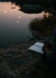 © CYBERPINK - Person's hand holding a pen and writing in an open notebook. the notebook is resting on a large rock, overlooking a body of water.