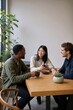 © Alex - Diverse group of friends chatting over coffee at a wooden table. Three people smiling and talking while holding mugs in a bright cafe