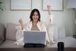 © matinee - Happy Asian woman celebrating success while using a laptop on her sofa at home.