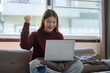 © matinee - Happy Asian woman celebrating success while using a laptop on her sofa at home.