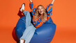 © pattozher - Joyful Indulgence: A vibrant young woman, radiating happiness, bounces on a large beanbag chair, arms outstretched in an expression of pure delight and carefree fun.