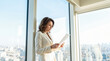 © Celt Studio - Caucasian businesswoman reading paper document standing beside large window modern office interior bright daytime city skyline background elegant white suit portrait