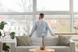 © Pixel-Shot - Young man behind sofa looking out large window at home, back view