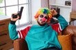 © Pixel-Shot - Young man dressed as crazy clown for Halloween with gun sitting on sofa in decorated living room