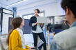 © saksit - Caucasian businessman holding laptop and presenting software updates to seated team members in office.
