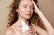 © SHOTPRIME STUDIO - Authentic beauty of a middle aged woman applying moisturizer on her cheek, close up on a neutral background, skincare, wellness, self care, natural look, unretouched