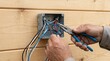 © NaesyaStudio - Electrician hands using pliers to connect wires in an electrical box mounted on a wooden wall