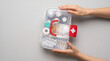 © Yuliani - Hands holding a portable first aid kit with medical supplies and medicine on a neutral grey surface