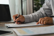 © onephoto - Business person reviewing a printed document with a pen on a clipboard beside a laptop, showing an office document checking process for paperwork verification and administrative review.