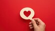 © MstKhadiza - A hand holding a speech bubble cutout with a red heart symbol inside, positioned against a solid, bright red background, minimalist concept