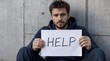 © Reandz - Young Sad Man Sitting Against Concrete Wall Holding White Help Sign, Social Issue Representation for Poverty