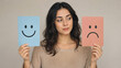 © Yuliia Yevdokymova - Young woman holding happy smiley and sad face cards choosing emotion on neutral background for mental health, customer feedback, mood swing