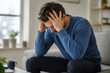 © aura - A stressed man sits on a couch holding his head in his hands, looking anxious and overwhelmed.
