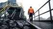 © Abode stock - Miner in high visibility jacket walking near conveyor belt system with coal pouring down at industrial facility during overcast day showing bulk raw material.