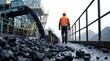 © Abode stock - Miner in high visibility jacket walking near conveyor belt system with coal pouring down at industrial facility during overcast day showing bulk raw material.