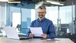 © Choirul - Professional middle-aged man with glasses reading documents in a modern office workspace.
