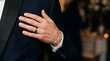 © wibiwisnu - Elegant man hand with wedding ring on dark tuxedo chest, focus on fabric texture and form, soft event lighting, rich negative space.