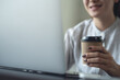 © tippapatt - Asian woman working remotely using laptop, hand holding a coffee cup, work at home, business casual, freelance lifestyle concept