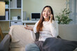 © matinee - Happy young Asian woman talking on a smartphone while relaxing with coffee at home.