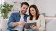 © Vyatcheslav - Happy young couple using digital tablet on sofa. Smiling man and woman looking at screen in modern living room. Online shopping and entertainment concept