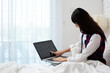 © Meng_Dakara - Woman touching laptop screen while working on bed with small white dog, remote work, home, technology, pet companionship, focus, comfort and digital lifestyle