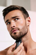 © Vitalii - Young man trimming the beard under his chin with an electric shaver, representing shaving, personal care, and grooming habits