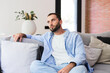 © Daniel - A bearded young man sits on a comfortable sofa in a bright modern living room, looking thoughtful and relaxed. Natural light, soft cushions and casual clothing create a calm, contemplative mood.