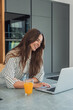 © Daniel - Young woman leans on a kitchen counter, smiling while using her laptop. Casual morning scene suggesting remote work, light breakfast and relaxed productivity in a modern home environment.