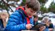 © Maksym Tereshchenko - A child at a summer camp under a heavy overcast sky peers at a small rugged tablet displaying animated malware detection graphics during an outdoor digital safety workshop, pine trees surrounding th