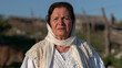 © InfinityVisuals - Elderly Woman in Traditional Attire: A weathered, elderly woman with a serene expression wears traditional white clothing and a headscarf, standing in a rustic outdoor setting under a clear sky.
