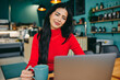 © Flamingo Images - Smiling woman in red sweater using laptop at a coffee shop with mug for remote work and productivity