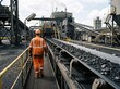 © mdnazirhossain - Industrial worker in orange reflective uniform walking along a coal conveyor belt at a large power plant facility
