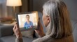 © Geetha - Over the shoulder view of senior woman watching virtual doctor on tablet, elderly patient engaged in telehealth consultation, glowing screen in modern living room