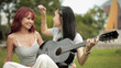 © vittaya - Smiling Asian Lesbian Couple Playing Guitar and Touching Hair in Green Park