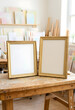 © Pixel-Shot - Art studio creativity and blank canvas mockup, two empty gold frames on a rustic wooden table with paintings