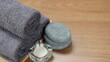 © sukanya - A minimalist still image in a spa setting shows dark gray towels rolled up on a wooden tray, with scented candles and stones placed beside them, creating a relaxing spa atmosphere.