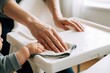 © Maryna - Mother cleans baby highchair tray while child grabs cloth. Parent wipes food mess, toddler interferes playfully. Daily home routine, bonding moment.